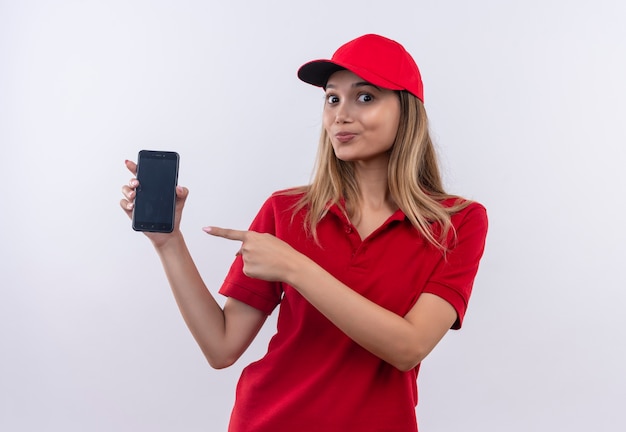 Zadowolona młoda dziewczyna dostawy ubrana w czerwony mundur i czapkę trzyma i wskazuje na telefon na białej ścianie