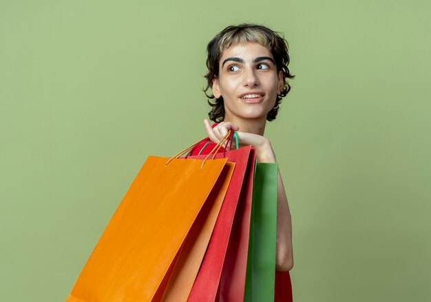 Zadowolona młoda dziewczyna kaukaska z fryzurą pixie, stojąca w widoku profilu, trzymająca torby na zakupy na ramieniu, patrząc za siebie odizolowana na oliwkowym tle z miejscem na kopię