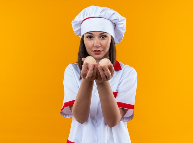 Zadowolona młoda kobieta kucharz ubrana w mundur szefa kuchni trzymająca jajka w aparacie na białym tle na pomarańczowym tle