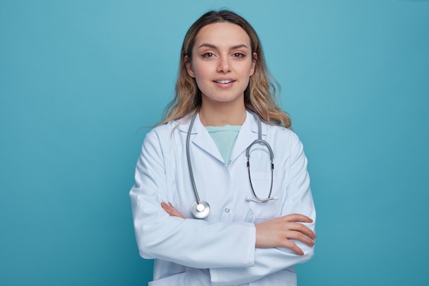 Zadowolona młoda kobieta lekarz ubrana w szlafrok medyczny i stetoskop wokół szyi, stojąca z zamkniętą posturą