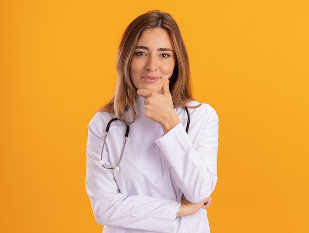Zadowolona młoda kobieta lekarz ubrana w szlafrok medyczny ze stetoskopem chwyciła brodę na białym tle na żółtej ścianie