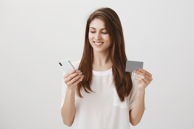 Zadowolona młoda kobieta robi zakupy online, pokazując kartę kredytową i telefon komórkowy