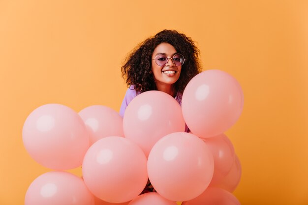 Zadowolona młoda kobieta w okrągłych okularach z różowymi balonami. radosne afrykańskie urodziny dziewczyna na żółtym tle.
