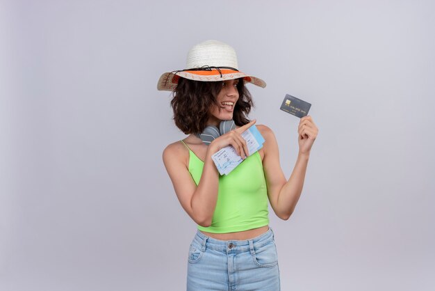 Zadowolona młoda kobieta z krótkimi włosami w zielonej bluzce w słuchawkach w kapeluszu przeciwsłonecznym trzymająca bilety lotnicze wskazując palcem wskazującym na kartę kredytową na białym tle