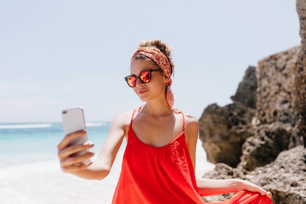 Zadowolona Młoda Kobieta Z Wstążką Włosów Dokonywanie Selfie Na Wybrzeżu Oceanu. Odkryty Zdjęcie Szczęśliwa Biała Dziewczyna Robi Sobie Zdjęcie Na Plaży.