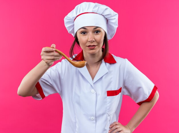 Zadowolona młoda kucharka ubrana w mundur szefa kuchni trzymająca łyżkę kładącą rękę na biodrze na białym tle na różowym tle