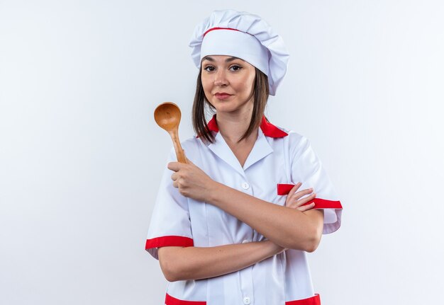 zadowolona młoda kucharka ubrana w mundur szefa kuchni trzymająca łyżkę krzyżującą ręce odizolowane na białej ścianie