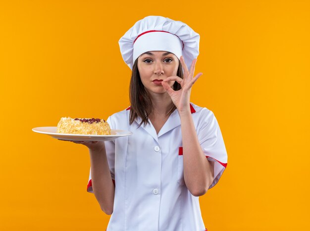 zadowolona młoda kucharka w mundurze szefa kuchni trzymająca ciasto na talerzu pokazującym pyszny gest na pomarańczowej ścianie!