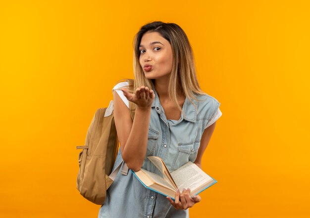 Zadowolona młoda ładna studencka dziewczyna ubrana w tylną torbę, trzymając otwartą książkę i wysyłając pocałunek ciosu z przodu na białym tle na pomarańczowej ścianie