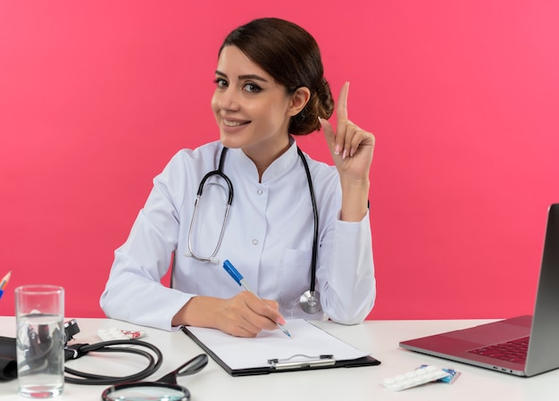 Zadowolona młoda lekarka w szlafroku medycznym ze stetoskopem siedząca przy biurku przy komputerze z narzędziami medycznymi napisz coś w schowku wskazuje na różową ścianę