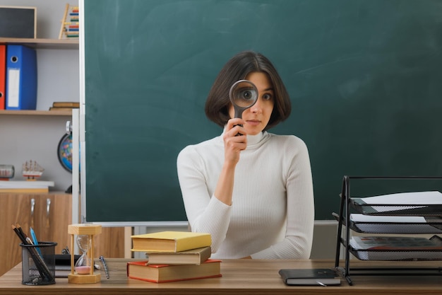 zadowolona młoda nauczycielka patrząca na kamerę z lupą siedzącą przy biurku z włączonymi narzędziami szkolnymi w klasie