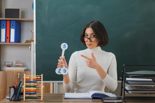 zadowolona młoda nauczycielka w okularach, trzymająca i wskazująca na wentylator numer, siedząca przy biurku z włączonymi narzędziami szkolnymi w klasie