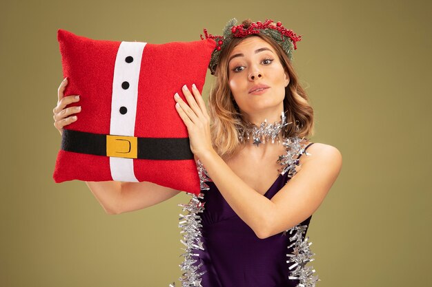 Zadowolona młoda piękna dziewczyna ubrana w fioletową sukienkę i wieniec z girlandą na szyi, trzymająca świąteczną poduszkę odizolowaną na oliwkowozielonym tle