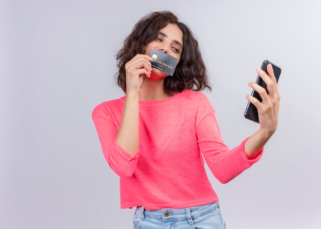 Zadowolona młoda piękna kobieta trzymając telefon komórkowy i kładąc kartę na ustach na odosobnionej białej ścianie