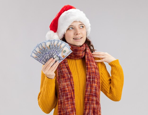 Zadowolona młoda słowiańska dziewczyna z santa hat i szalikiem na szyi trzymająca pieniądze i patrząca na bok odizolowaną na białej ścianie z kopią przestrzeni