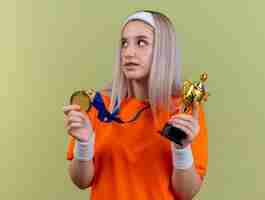 Bezpłatne zdjęcie zadowolona młoda sportowa dziewczyna rasy kaukaskiej z szelkami w opasce i opaskach na nadgarstku trzyma złoty medal i puchar zwycięzcy patrząc na bok