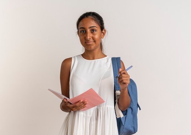Zadowolona młoda uczennica na sobie tylną torbę, trzymając notatnik i długopis na białym tle