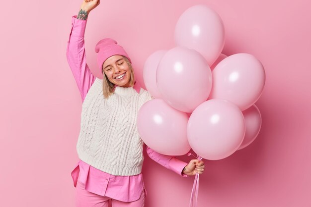 Zadowolona, optymistyczna kobieta nosi modne ubrania tańczy do muzyki na imprezie ma świąteczny nastrój pozuje na różowym tle z napompowanymi balonami świętuje urodziny z przyjaciółmi Koncepcja wakacji