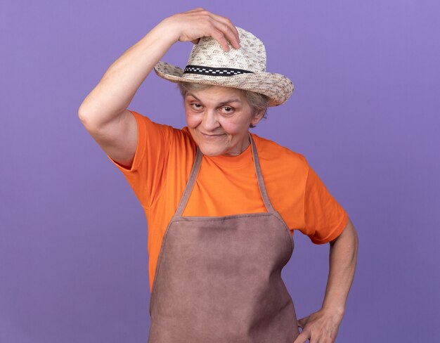Zadowolona starsza ogrodniczka w kapeluszu ogrodniczym kładzie rękę na kapeluszu na fioletowo