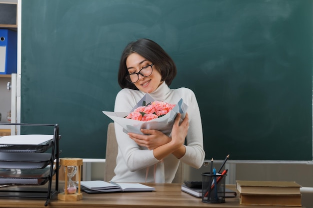 zadowolona z zamkniętych oczu młoda nauczycielka siedzi przy biurku ze szkolnymi narzędziami i trzyma bukiet kwiatów w klasie