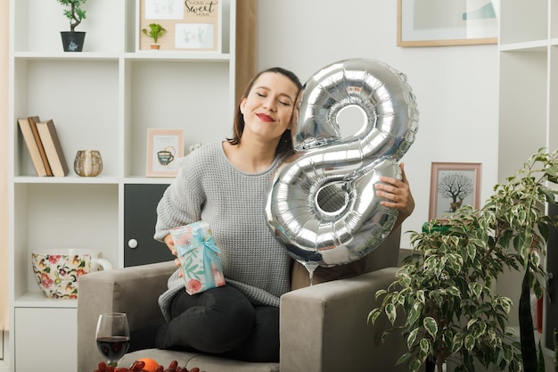 Zadowolona z zamkniętych oczu piękna kobieta w szczęśliwy dzień kobiet trzymająca balon z numerem osiem z prezentem siedzącym na fotelu w salonie