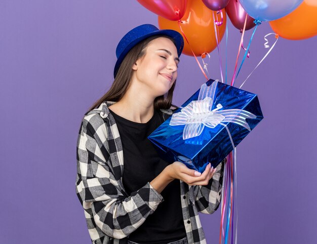 Zadowolona z zamkniętymi oczami młoda piękna dziewczyna w kapeluszu imprezowym trzymająca balony z pudełkiem na prezent