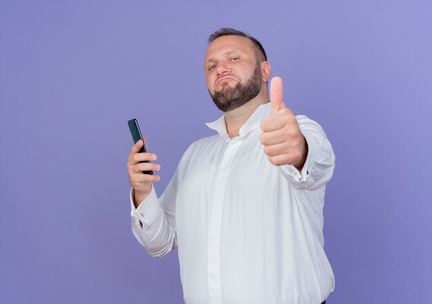 Bezpłatne zdjęcie zadowolony brodaty mężczyzna ubrany w białą koszulę, trzymając smartfona, uśmiechnięty i pewny siebie, pokazując kciuki do góry stojąc nad niebieską ścianą