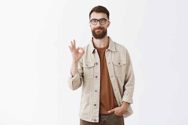 Zadowolony brodaty mężczyzna w okularach pozuje przy białej ścianie