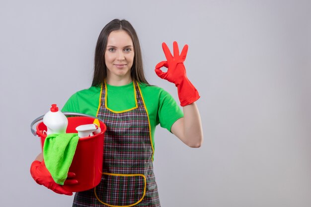 Zadowolony czyszczenia młoda dziewczyna ubrana w mundur w czerwonych rękawiczkach, trzymając narzędzia do czyszczenia, pokazując gest okey na na białym tle