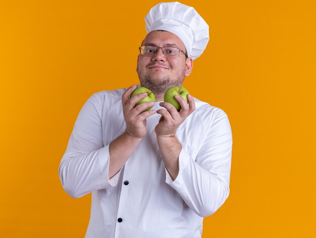 zadowolony dorosły mężczyzna kucharz ubrany w mundur szefa kuchni i okulary trzymający jabłka patrzący na przód odizolowany na pomarańczowej ścianie