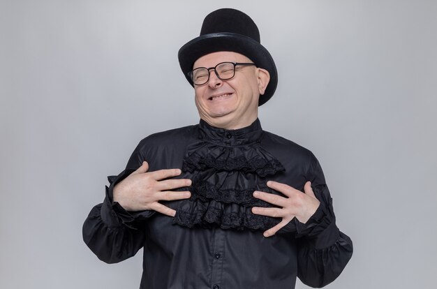 Zadowolony dorosły słowiański mężczyzna w cylindrze i okularach optycznych w czarnej gotyckiej koszuli, kładąc ręce na jego klatce piersiowej