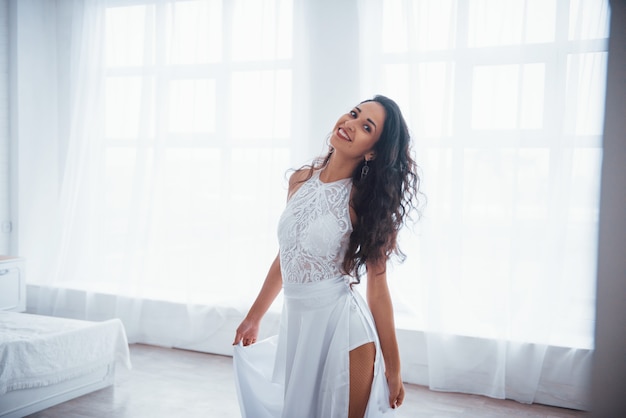 Zadowolony i bezpłatny. Piękna kobieta w białej sukni stoi w białym pokoju ze światłem dziennym przez okna