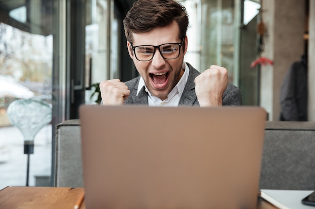 Zadowolony krzyczący biznesmen siedzi przy stoliku w kawiarni w okularach i raduje się, patrząc na laptopa