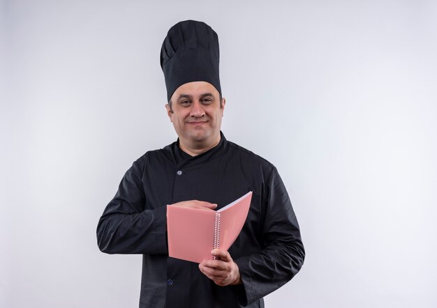 Zadowolony kucharz w średnim wieku w mundurze szefa kuchni trzymając notebook na odosobnionej białej ścianie z miejscem kopiowania