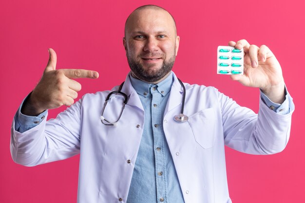 Bezpłatne zdjęcie zadowolony lekarz w średnim wieku ubrany w szatę medyczną i stetoskop pokazujący opakowanie kapsułek medycznych do kamery wskazującej na to izolowane na różowej ścianie