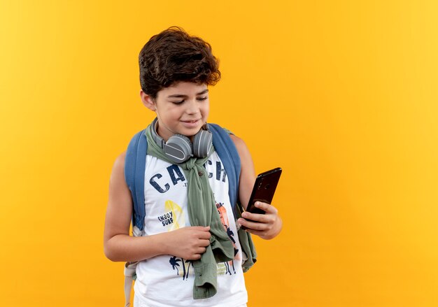Zadowolony mały chłopiec w szkole noszenia plecaka i słuchawek, trzymając i patrząc na telefon na białym tle na żółtym tle