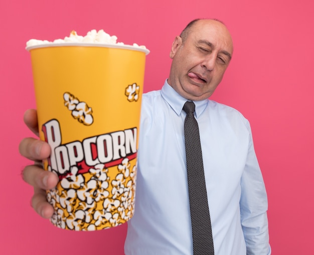 Zadowolony mężczyzna w średnim wieku, ubrany w białą koszulkę z krawatem, trzymający wiadro popcornu w aparacie odizolowany na różowej ścianie