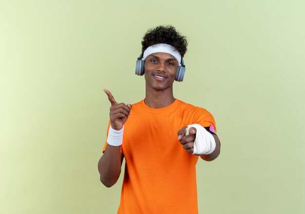 Zadowolony młody afro-amerykański sportowy mężczyzna z opaską na głowę i opaską na rękę i opaską na ramię telefonu ze słuchawkami pokazującymi gest i punkty z boku na białym tle na zielonym tle z miejscem na kopię