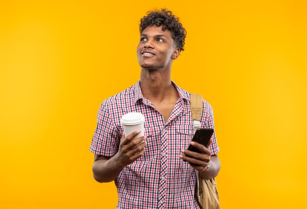 Zadowolony młody afroamerykański student z plecakiem trzymającym telefon i papierowy kubek patrzący na bok