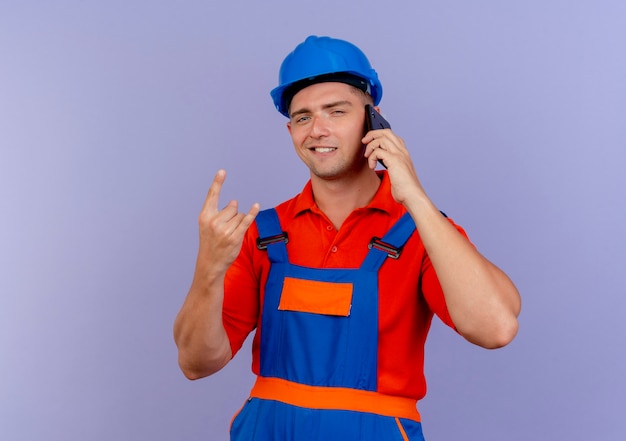 Zadowolony młody budowniczy mężczyzna w mundurze i kasku ochronnym rozmawia przez telefon i pokazuje gest kozy na fioletowo