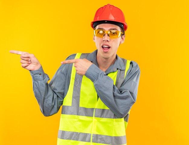 Zadowolony młody budowniczy mężczyzna w mundurze w okularach wskazuje z boku na żółtej ścianie z miejscem na kopię