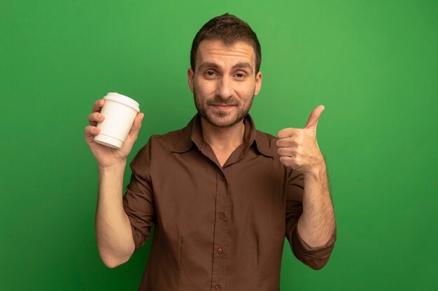 Zadowolony młody człowiek patrząc z przodu trzymając plastikowy kubek do kawy pokazując kciuk do góry na białym tle na zielonej ścianie