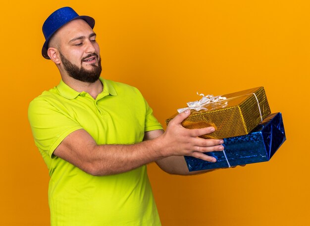 Zadowolony młody człowiek w imprezowym kapeluszu trzymającym pudełka z prezentami z boku