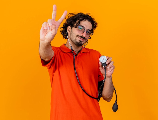 Zadowolony młody kaukaski chory mężczyzna w okularach i stetoskopie trzymający ciśnieniomierz robi znak pokoju na pomarańczowej ścianie z miejscem na kopię