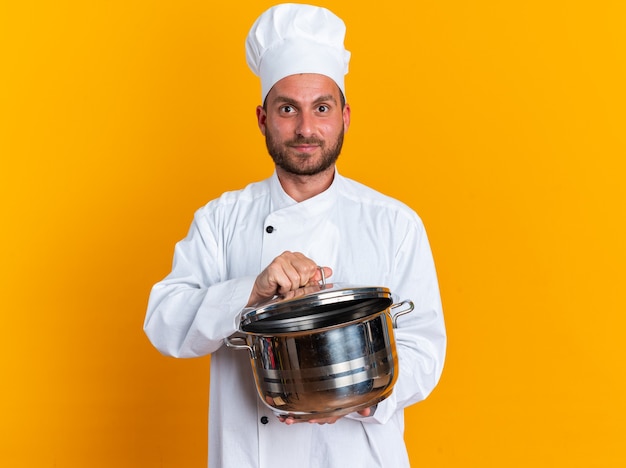 Zadowolony młody kaukaski kucharz w mundurze szefa kuchni i czapce trzymającej garnek chwytający pokrywkę garnka