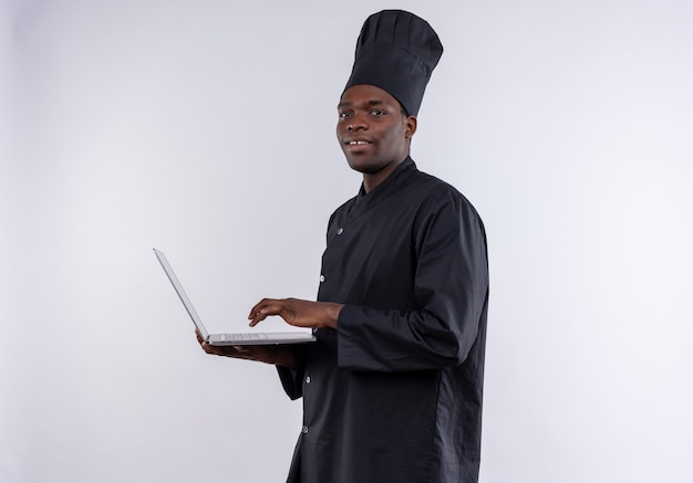 Zadowolony młody kucharz afro-amerykański w mundurze szefa kuchni trzyma laptopa i patrzy na aparat na białym z miejsca na kopię
