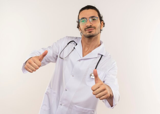 Zadowolony młody lekarz mężczyzna z okularami optycznymi na sobie białą szatę ze stetoskopem kciuki do góry na odosobnionej białej ścianie z miejsca na kopię