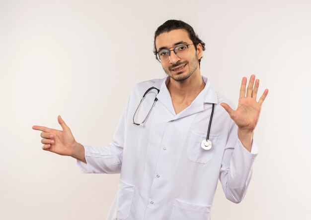 Zadowolony młody lekarz płci męskiej z okularami optycznymi w białej szacie ze stetoskopem pokazującym gest zatrzymania i wskazujący na bok na odizolowanej białej ścianie z miejscem na kopię