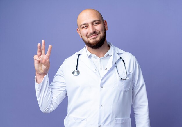 Zadowolony młody łysy lekarz mężczyzna ubrany w szlafrok i stetoskop pokazujący trzy na białym tle na niebieskim tle