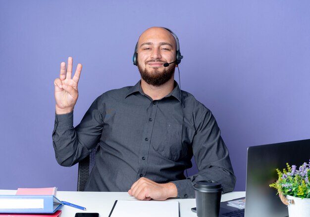 Bezpłatne zdjęcie zadowolony młody łysy mężczyzna call center sobie zestaw słuchawkowy siedzi przy biurku z narzędzi pracy pokazując trzy na białym tle na fioletowym tle